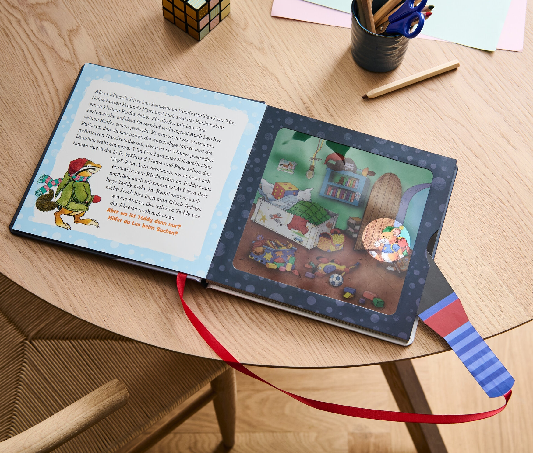 Das Buch "Leo Lausemaus – mein Taschenlampenbuch" liegt geöffnet auf einem Tisch neben Buntstiften und einem Würfel.