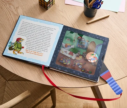 Das Buch "Leo Lausemaus – mein Taschenlampenbuch" liegt geöffnet auf einem Tisch neben Buntstiften und einem Würfel.