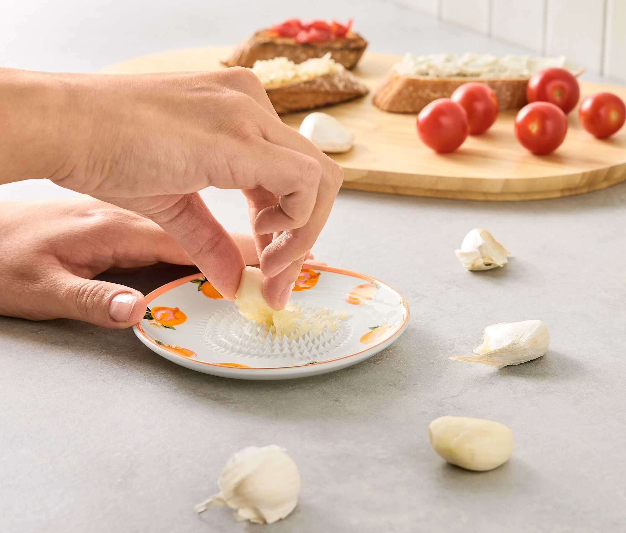Eine Person reibt Knoblauch auf einer Tellerreibe. Im Hintergrund sind Toasts und Tomaten.