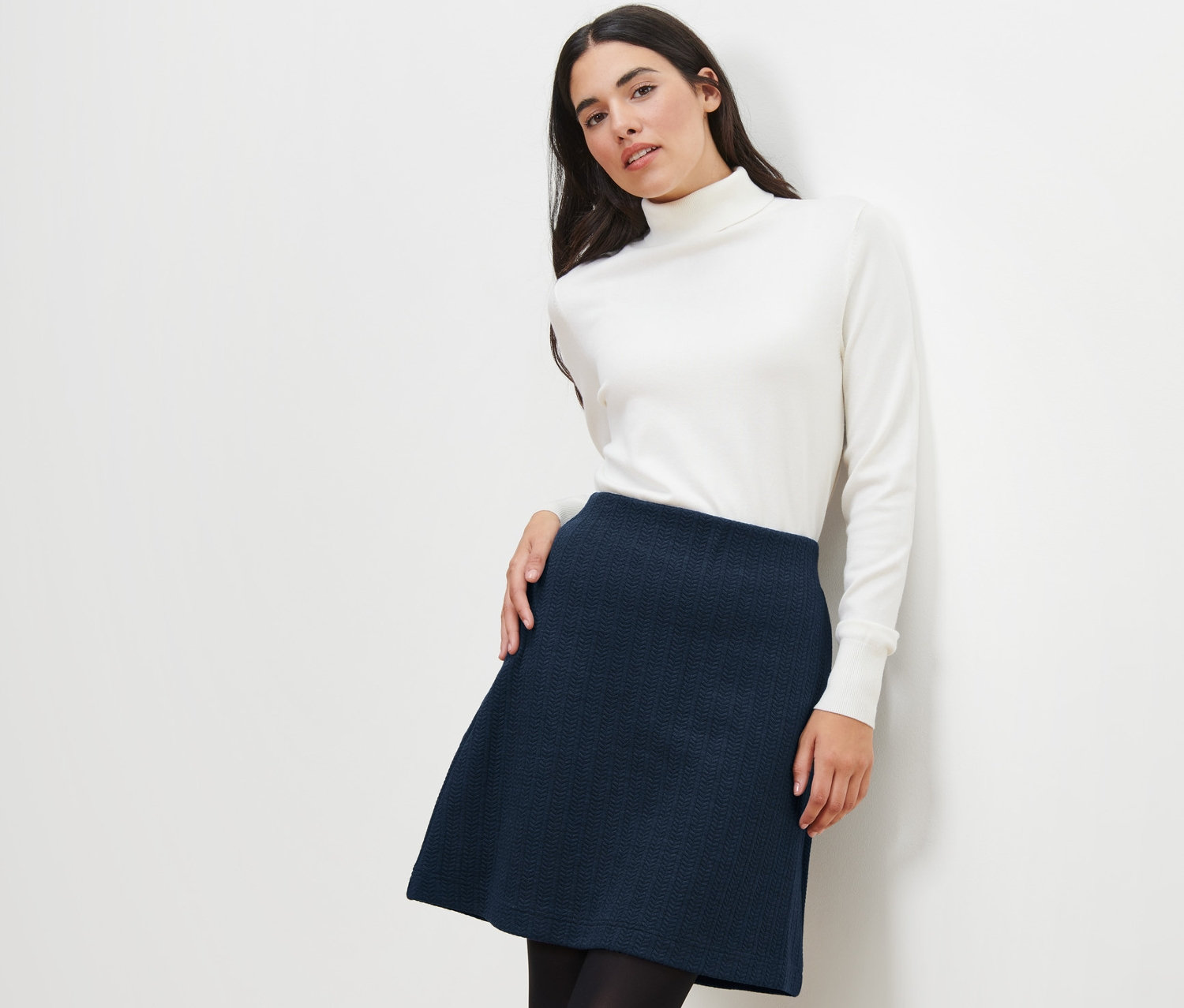 Frau posiert in einem Sweatrock mit Zopfmuster und einem weißen Rollkragenpullover.