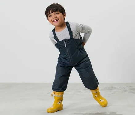 Ein Junge posiert in einer Kinder-Regenhose mit Mitwachsfunktion und gelben Gummistiefeln.