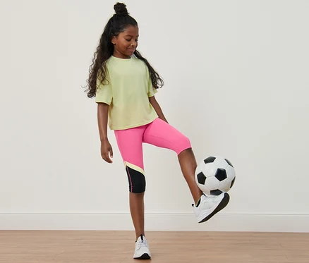 Ein Mädchen spielt mit einem Fußball, sie trägt eine 2 Kinder-Sporttights, Radler- und 3/4-Länge.