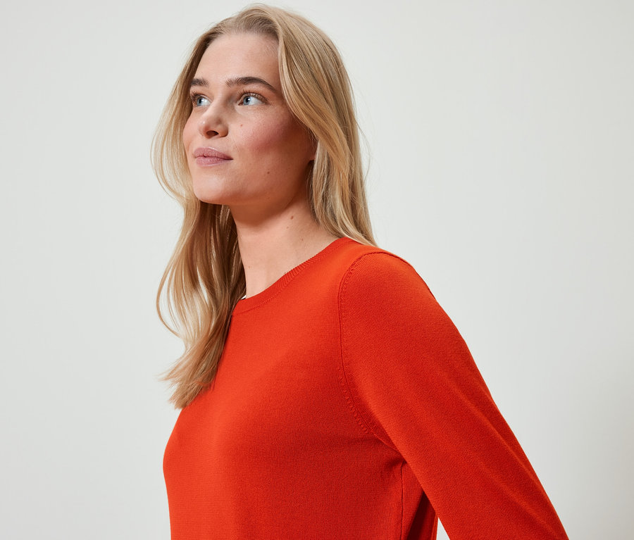 Porträt einer Frau mit blonden Haaren in einem orangefarbenen Pullover, die nach links schaut.
