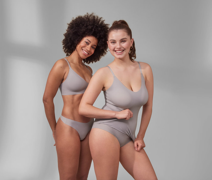 Zwei lächelnde Frauen posieren im Studio. Eine trägt einen Komfort-BH »bonded« und 5 Magic-Cut-Strings. Die andere trägt ein Komfort-Hemdchen »bonded« und 5 Magic-Cut-Pantys.