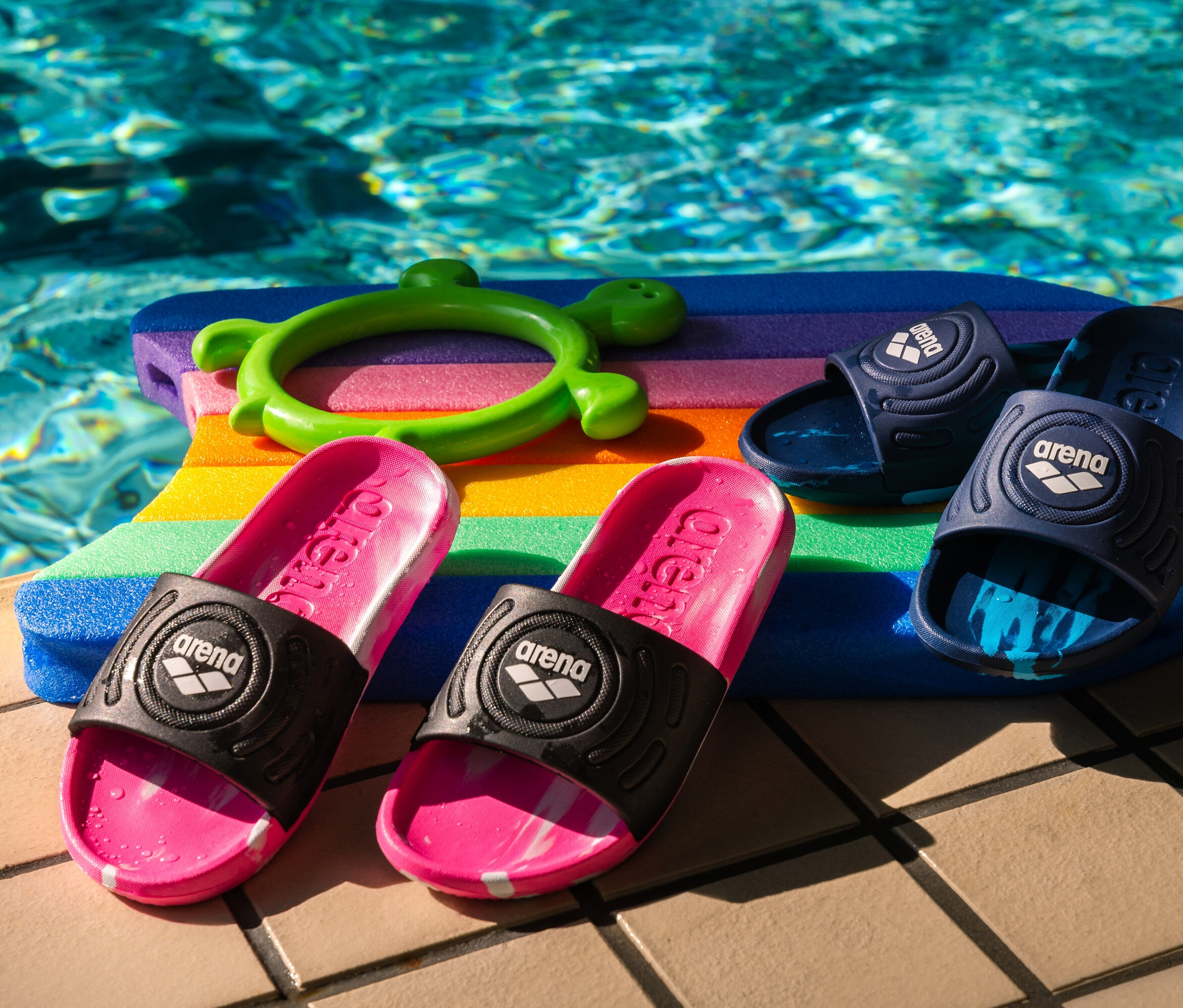 Zwei Paar arena Kinder-Badesandalen Junior Urban Active, pink und navy, liegen auf einem bunten Schwimmbrett am Pool.