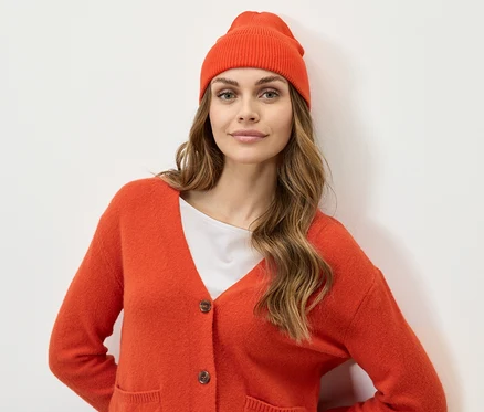 Frau mit braunen Haaren, orangefarbener Mütze und Strickjacke blickt in die Kamera.