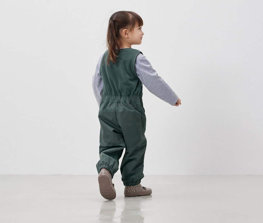 Mädchen in grüner Latzhose und grauem Hemd geht auf einem weißen Boden.