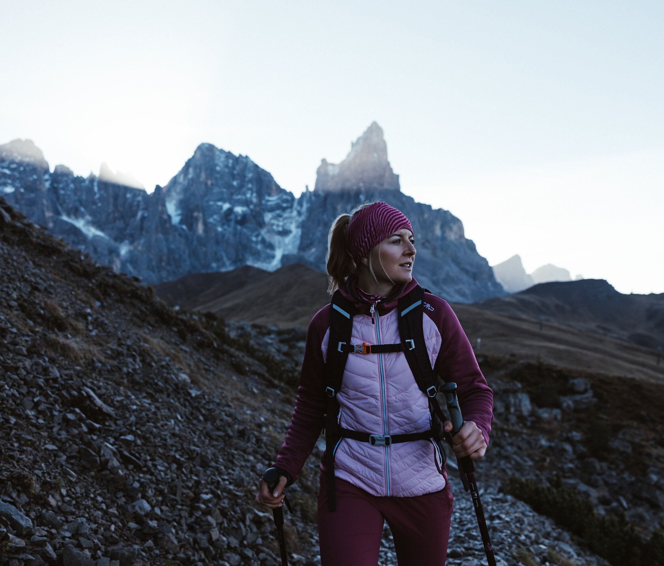 Eine Frau mit Rucksack und Trekkingstöcken wandert bergauf. Sie trägt eine CMP Damen-Hybridjacke, beere.