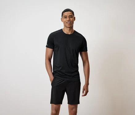 Ein lächelnder Mann steht mit den Händen in den Taschen seiner schwarzen Shorts und trägt ein schwarzes T-Shirt.