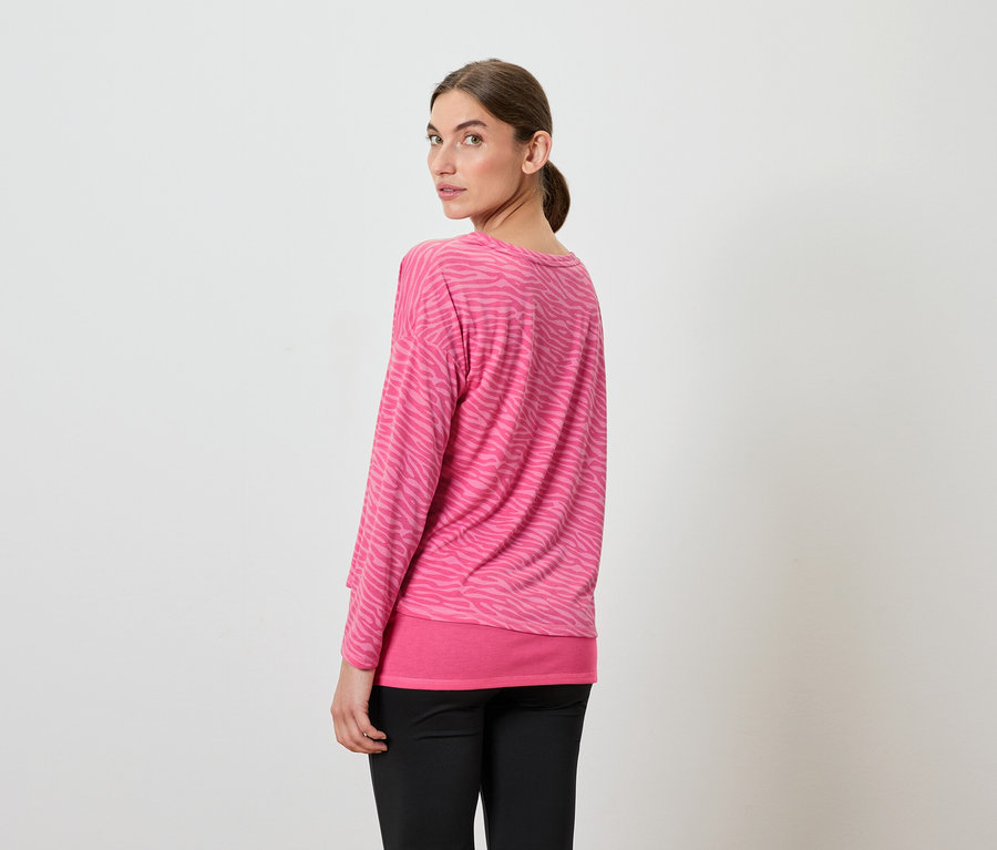 Frau blickt über die Schulter, trägt ein rosa Langarmshirt und eine schwarze Hose.