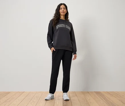 Frau steht mit Händen in den Taschen ihrer schwarzen Jogginghose. Sie trägt ein graues Sweatshirt mit der Aufschrift Sports Club und weiße Turnschuhe.