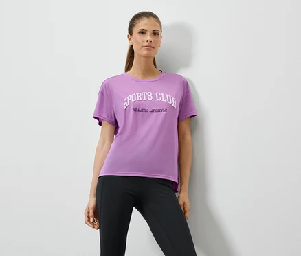 Frau in lila T-Shirt mit Aufdruck und schwarzen Leggings posiert vor einer weißen Wand.