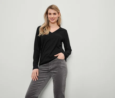 Blonde Frau in schwarzem Langarmpullover und grauer Cordhose steht und lächelt.