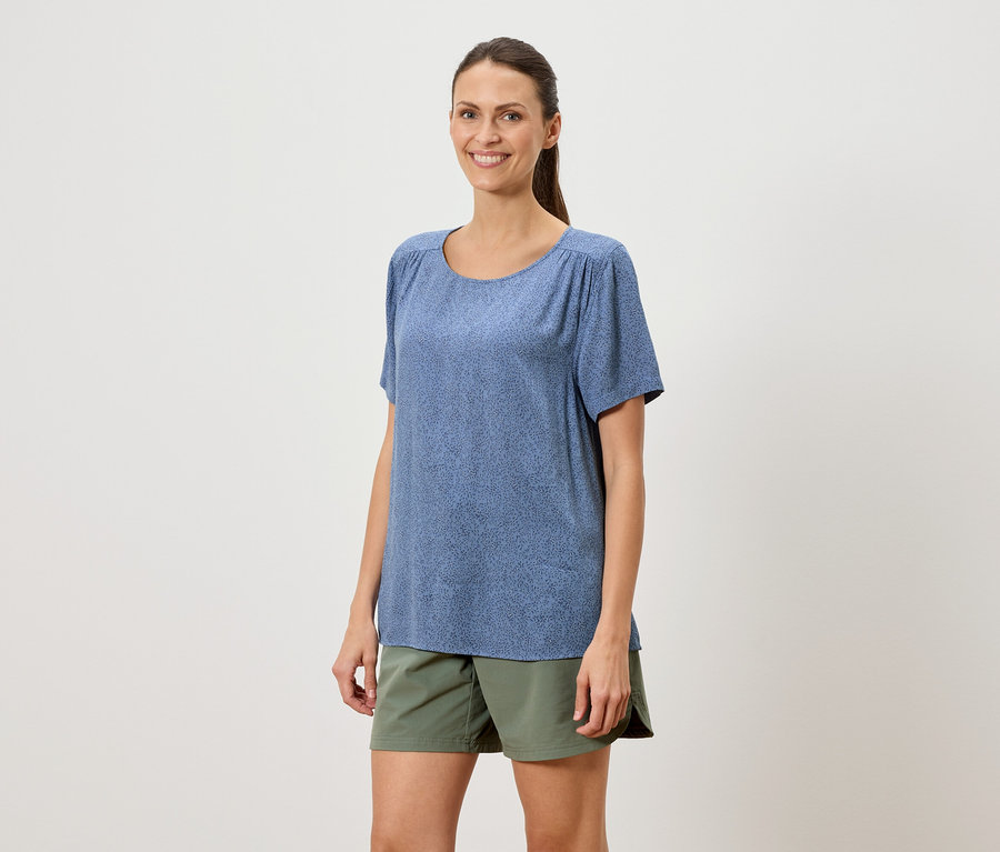 Frau mit Pferdeschwanz trägt ein blaues T-Shirt mit kurzem Arm und eine grüne Shorts vor einem weißen Hintergrund.