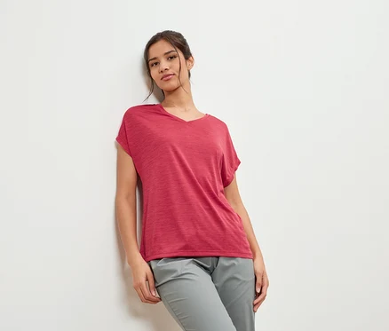 Junge Frau lehnt an einer Wand, sie trägt ein weinrotes T-Shirt und graue Hosen.
