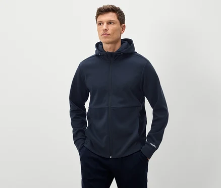 Mann posiert mit marineblauem Kapuzenpullover mit durchgehendem Reißverschluss und marineblauer Hose, die mit beiden Händen in den Taschen.