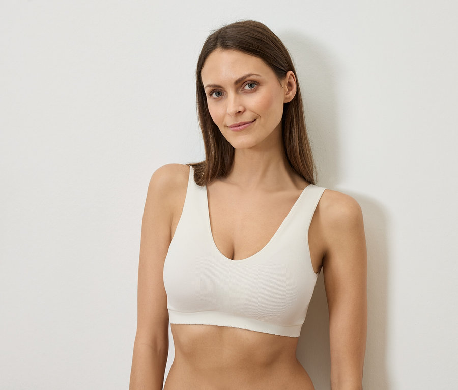 Frau mit langen braunen Haaren, die einen weißen BH trägt und gegen eine weiße Wand lehnt.