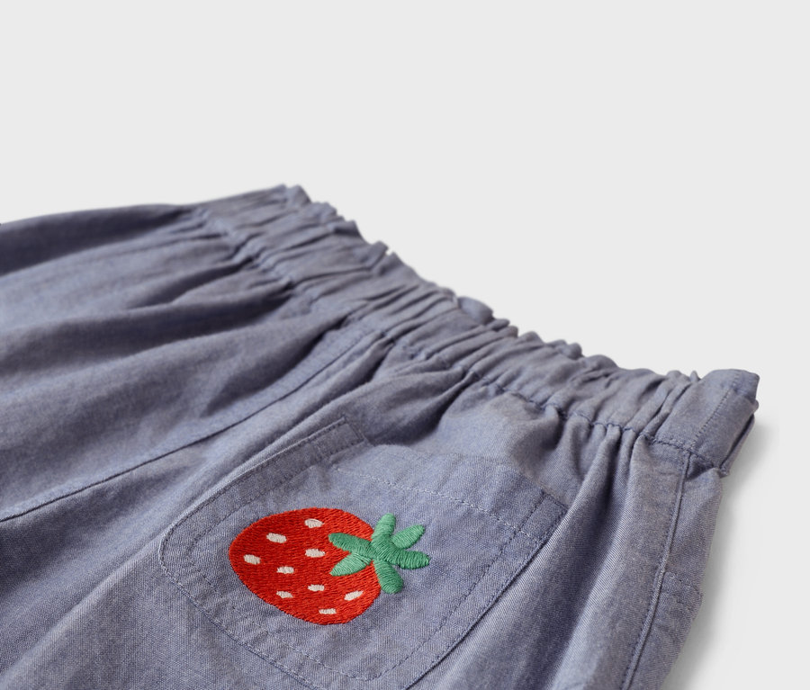 Nahaufnahme einer blauen Stoffhose mit Gummibund und einer aufgesetzten Tasche, auf der eine rote Erdbeere aufgestickt ist.