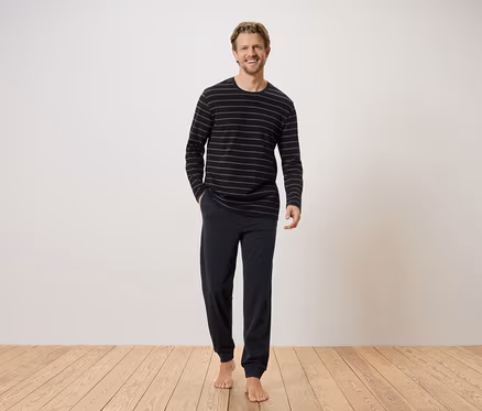 Mann mit blonden Haaren, der ein gestreiftes, langärmeliges Oberteil und eine dunkle Hose vor einer weißen Wand trägt.