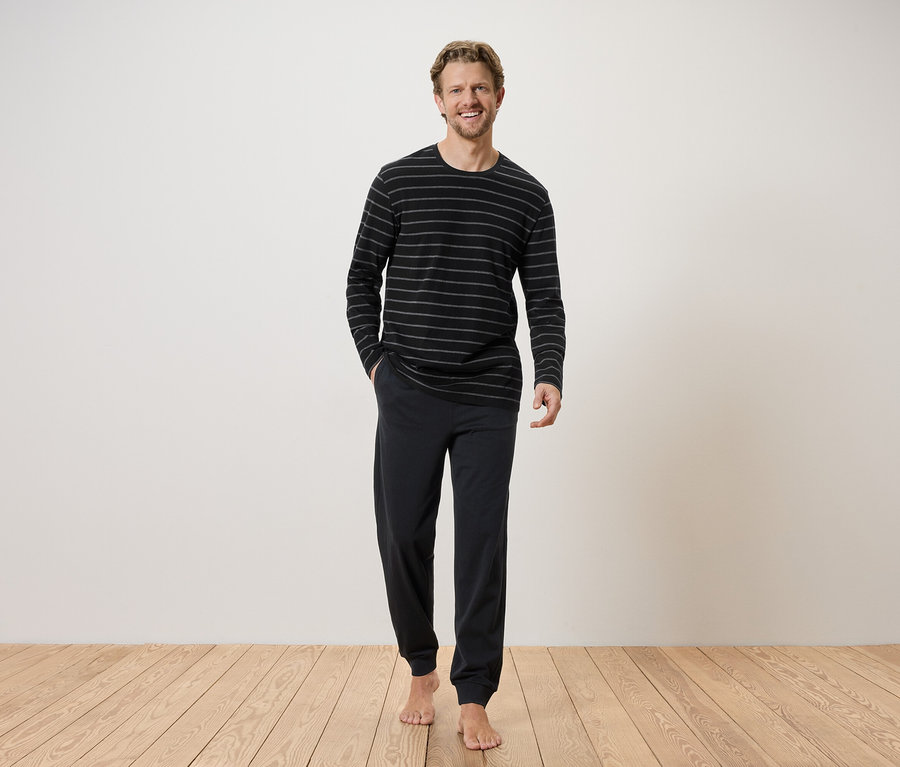 Mann mit blonden Haaren, der ein gestreiftes, langärmeliges Oberteil und eine dunkle Hose vor einer weißen Wand trägt.