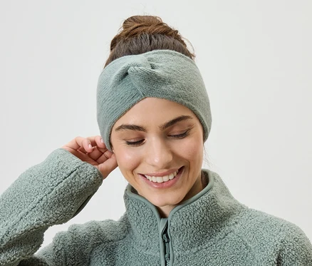 Frau mit hochgesteckten Haaren und Stirnband in Hellgrün, trägt eine hellgrüne Fleecejacke und berührt ihr Ohr.