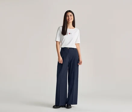 Eine Frau mit langen, dunklen Haaren lächelt und trägt ein weißes T-Shirt mit drei roten Herzen und eine weite, marineblaue Hose.