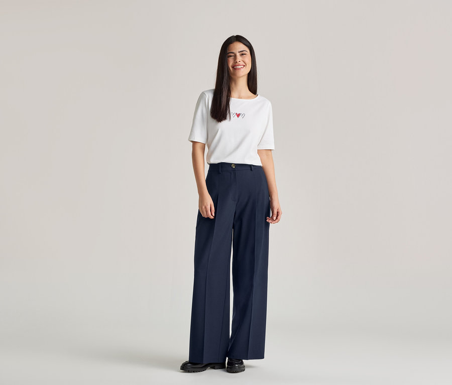 Eine Frau mit langen, dunklen Haaren lächelt und trägt ein weißes T-Shirt mit drei roten Herzen und eine weite, marineblaue Hose.