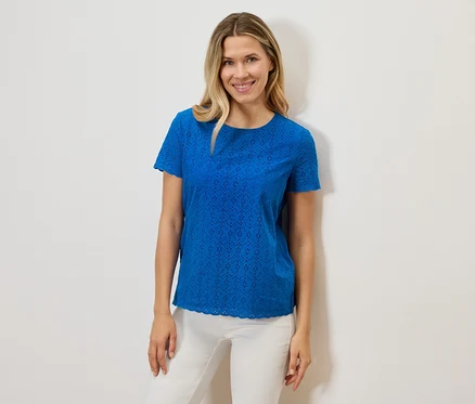 Eine Frau lächelt und trägt ein blaues, kurzärmeliges Lochmuster-T-Shirt und eine weiße Hose vor einem weißen Hintergrund.