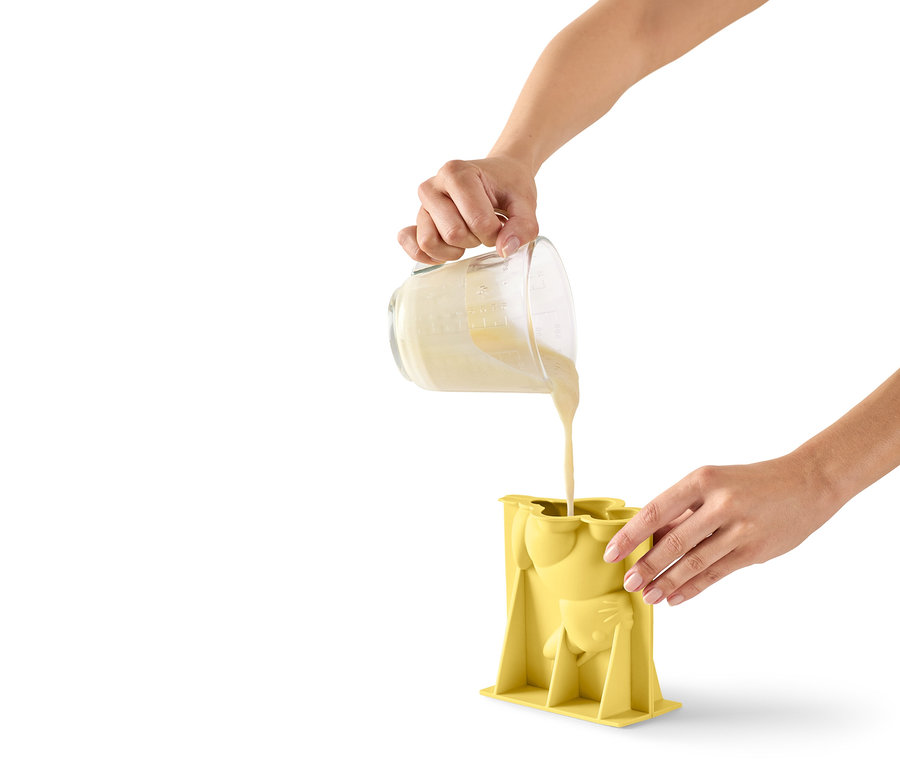 Eine Person gießt Teig aus einem Messbecher in eine gelbe, katzenförmige Silikonform.