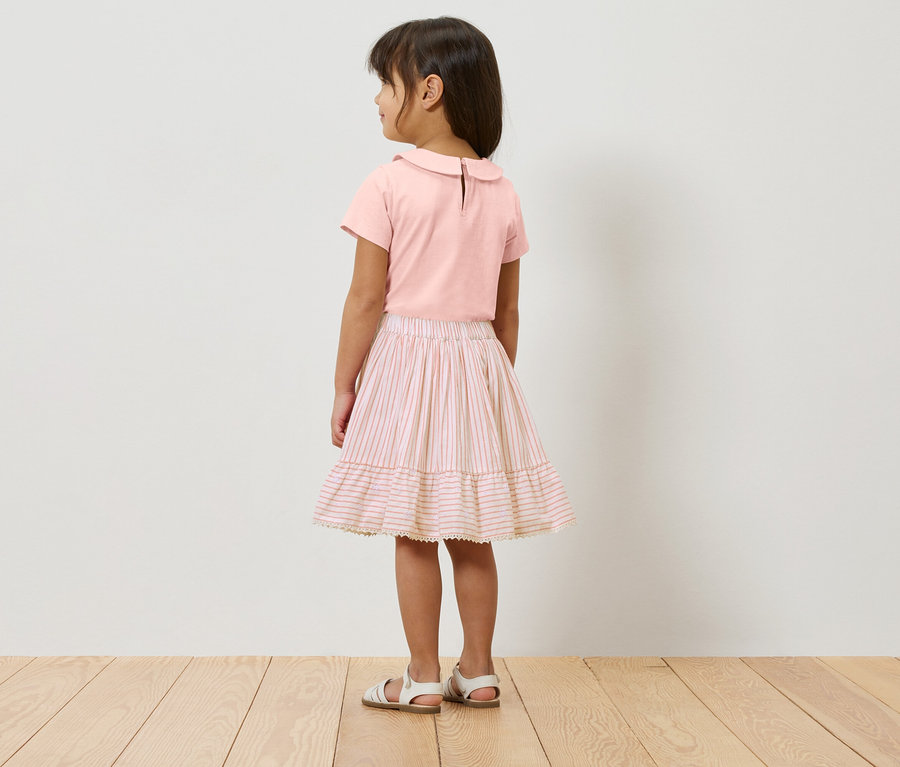 Ein Mädchen steht mit dem Rücken zur Kamera, trägt ein rosa Oberteil, einen rosa-weiss gestreiften Rock und weisse Sandalen.