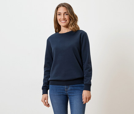 Eine Frau mit gewelltem, braunem Haar trägt einen marineblauen Pullover und Jeans vor einem weißen Hintergrund.