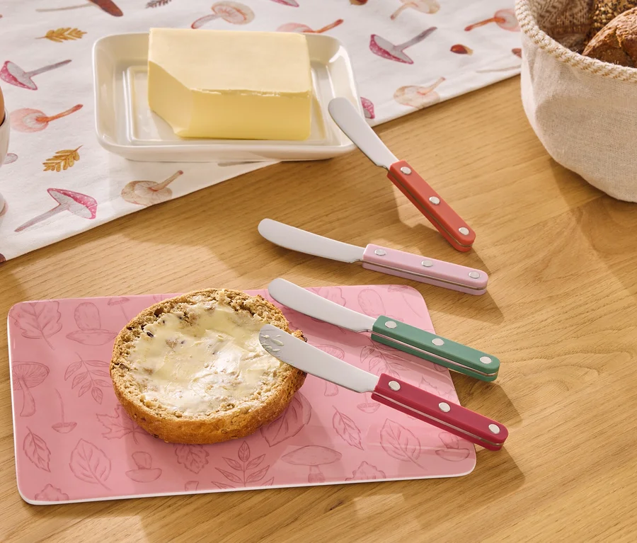 Auf einem Holztisch liegen ein Ei im Eierbecher, Butter, ein Brötchen mit Butter und vier Buttermesser in Rot, Rosa, Grün und Rot.