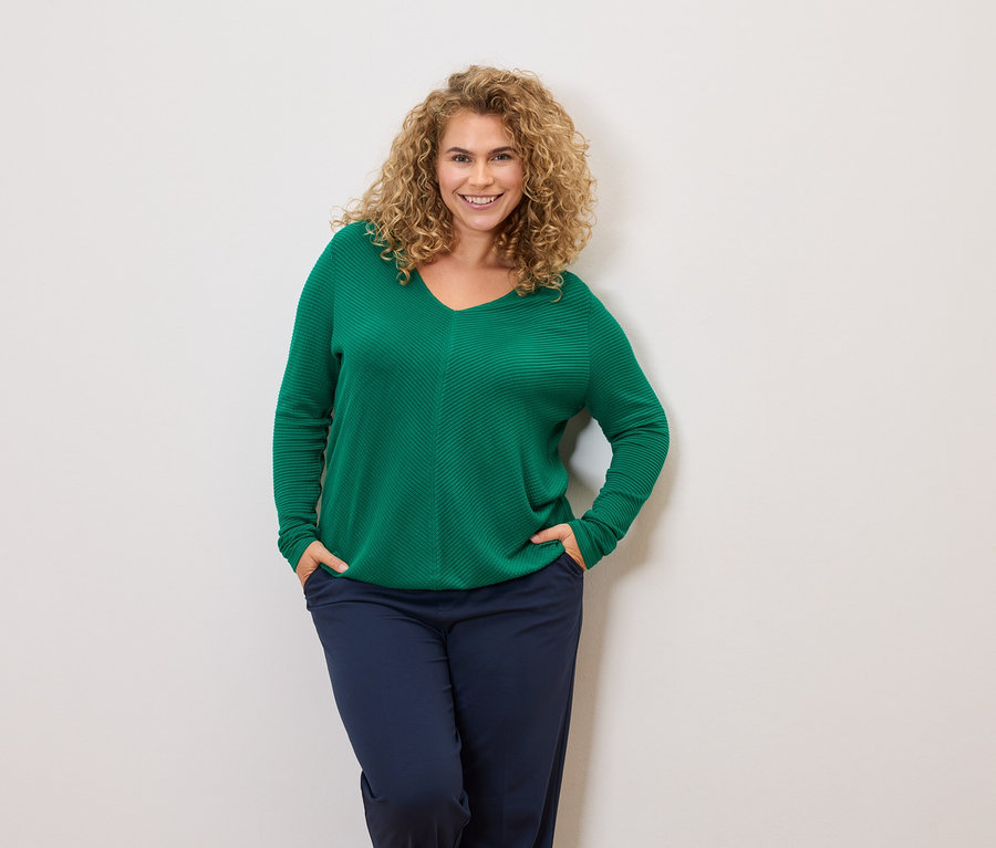 Frau mit lockigem Haar trägt einen grünen Pullover und marineblaue Hosen, die Hände in den Taschen.