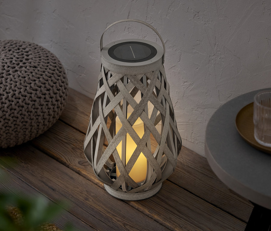 Eine geflochtene Laterne mit Solarpanel steht neben einem Pouf auf einem Holzboden.