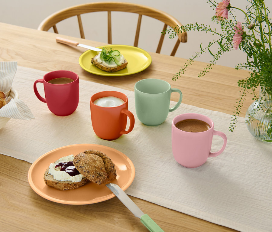 Holztisch mit hellem Tischläufer, vier bunten Tassen (rot, orange, mintgrün, rosa) mit Getränken, zwei Tellern mit belegten Brötchen, Brotkorb und Blumenvase.
