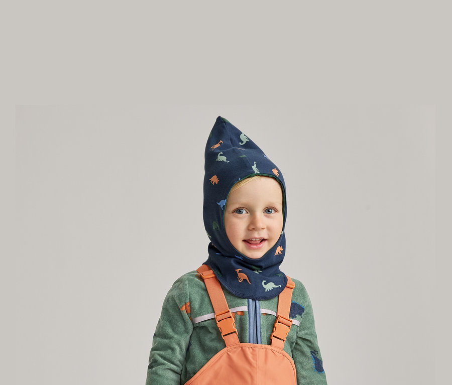 Lächelnder Junge mit blauem Dino-Balaclava, grünem Fleecepullover und orangefarbener Latzhose vor grauem Hintergrund.