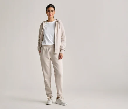 Frau mit Zopf trägt beiges Trainingsoutfit mit Kapuzenjacke, T-Shirt und Jogginghose, dazu weiße Turnschuhe.