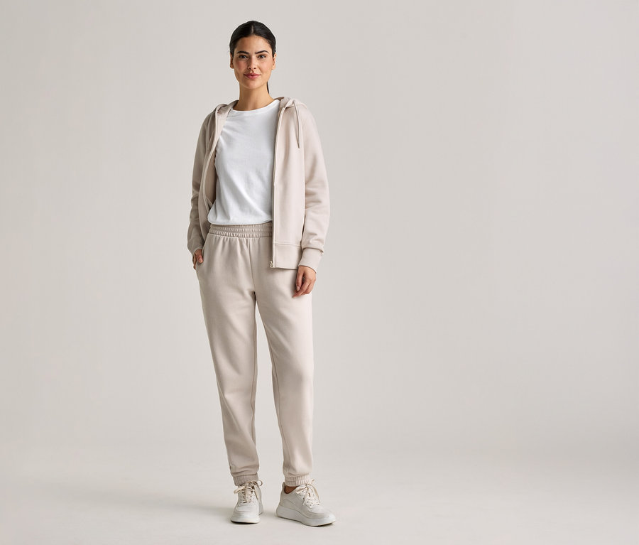 Frau mit Zopf trägt beiges Trainingsoutfit mit Kapuzenjacke, T-Shirt und Jogginghose, dazu weiße Turnschuhe.
