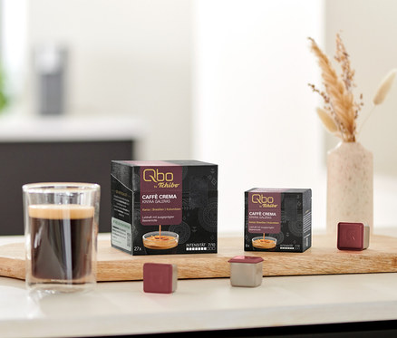 Ein Glas Kaffee steht neben Qbo Caffè KINYAA GALERAS Kapselpackungen auf einem Holzbrett.