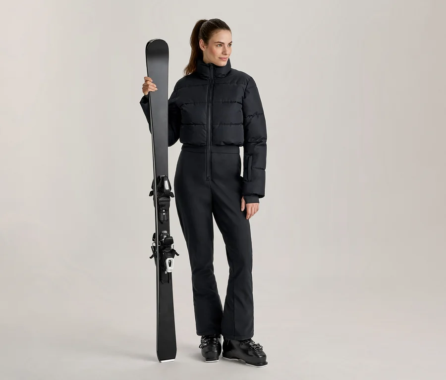 Frau in schwarzer, kurzer Daunen-Skijacke und schwarzer Skihose, die einen schwarzen Ski vertikal hält und schwarze Skischuhe trägt.