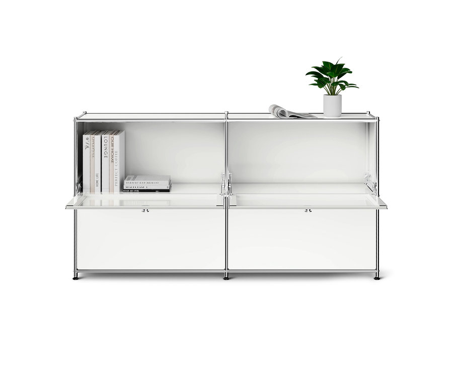 Weißes modulares Sideboard mit Chromrahmen. Die obere linke Ablage enthält Bücher, die obere rechte ist leer. Auf der rechten Seite stehen eine Pflanze und eine gerollte Zeitung. Die unteren Klapptüren sind leicht geöffnet.