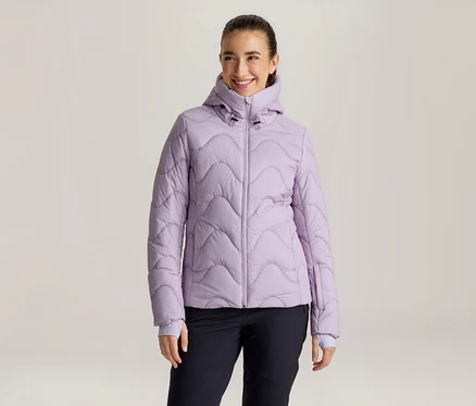 Lächelnde Frau mit zurückgebundenen dunklen Haaren, die eine hellviolette Steppjacke mit Wellenmuster und Kapuze sowie eine dunkle Hose trägt und nach rechts blickt.