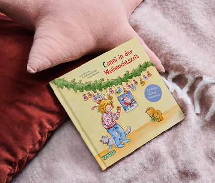 Das Buch Conni in der Weihnachtszeit liegt auf einer Decke neben rosa Kissen.