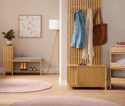Heller Vorraum mit einem Kleiderständer mit Ablage, an dem Jacken hängen. In der Mitte eine Sitzbank mit Schuhfach und Ablage in Lamellenoptik mit Kissen und Schuhen darunter, daneben eine Dreibein-Stehleuchte aus Holz. Rechts eine Garderobe mit Sitzbank in Lamellenoptik mit Jacke, Schal und Tasche. Daneben ein Standregal aus Massivholz mit 3 Fächern. Auf dem Boden liegen zwei pinke Teppiche „Charme“.