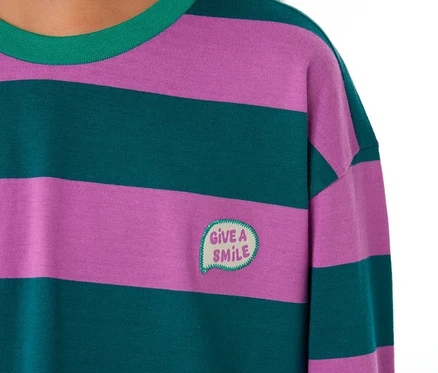 Nahaufnahme eines Kindes, das ein SCHIESSER Kinder-Pyjama Oberteil mit lila und dunkelgrünen Querstreifen, grünem Kragen und einem aufgestickten „GIVE A SMILE“-Sprechblasen-Patch trägt.