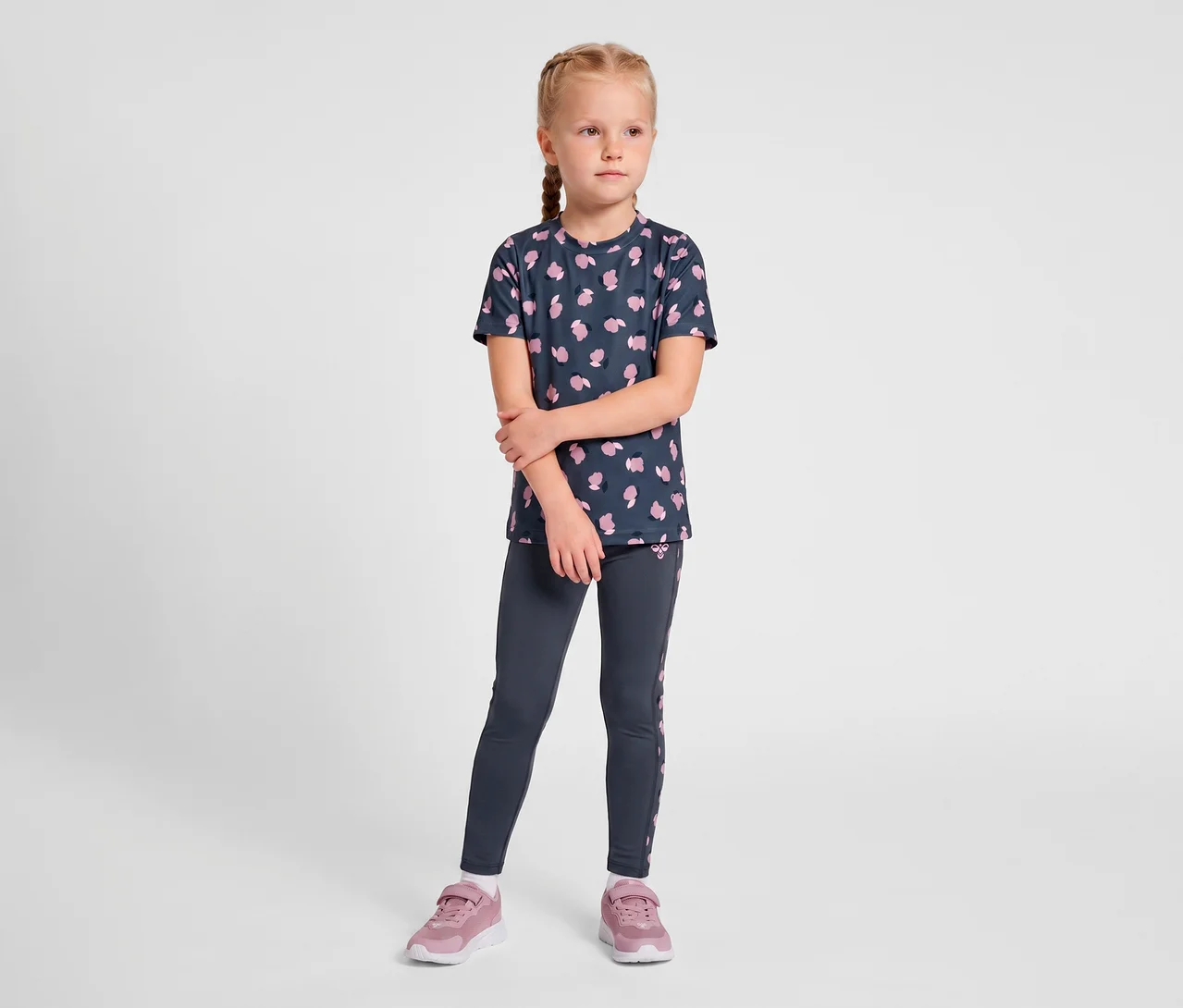 Mädchen in dunkelblauem T-Shirt mit pinkem Muster und dunkelgrauen hummel® »HMLJR AOP Gym Tights« mit pinken Seitenstreifen, dazu pinke Turnschuhe.