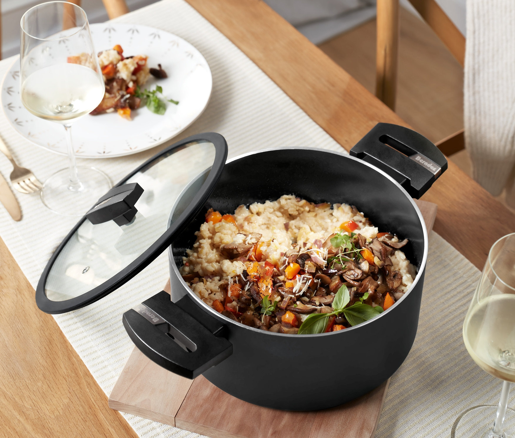 Auf einem Tisch steht ein schwarzer Kochtopf mit Glasdeckel »Alu Recycled Induction b.green«, gefüllt mit Risotto. Daneben ein Glas Wein und ein Teller mit Risotto.