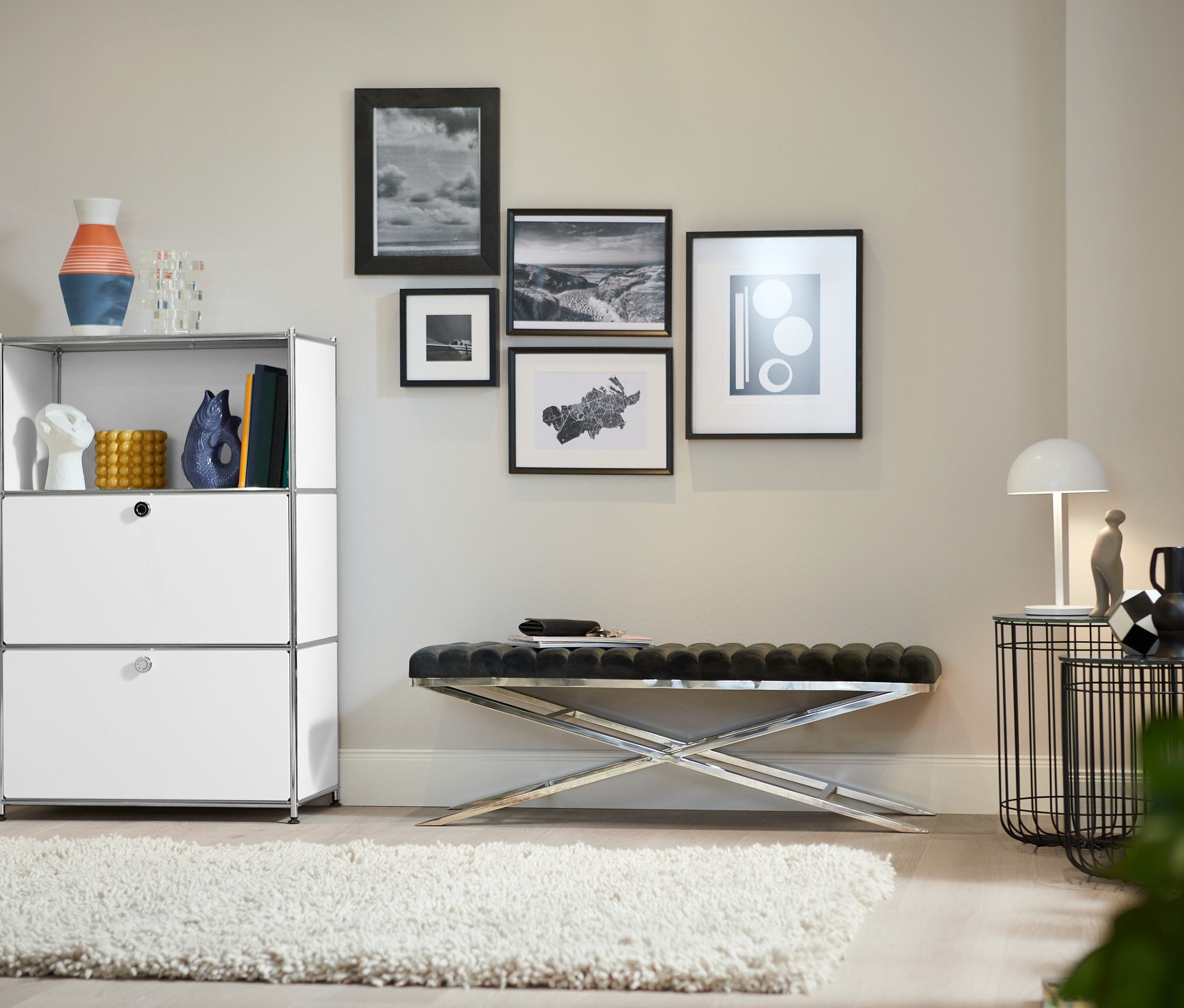 Ein Wohnzimmer zeigt ein Metallregal mit 2 weißen Klappenfächern, eine Design-Bank mit Metallgestell und einen Design-Barwagen aus Metall.