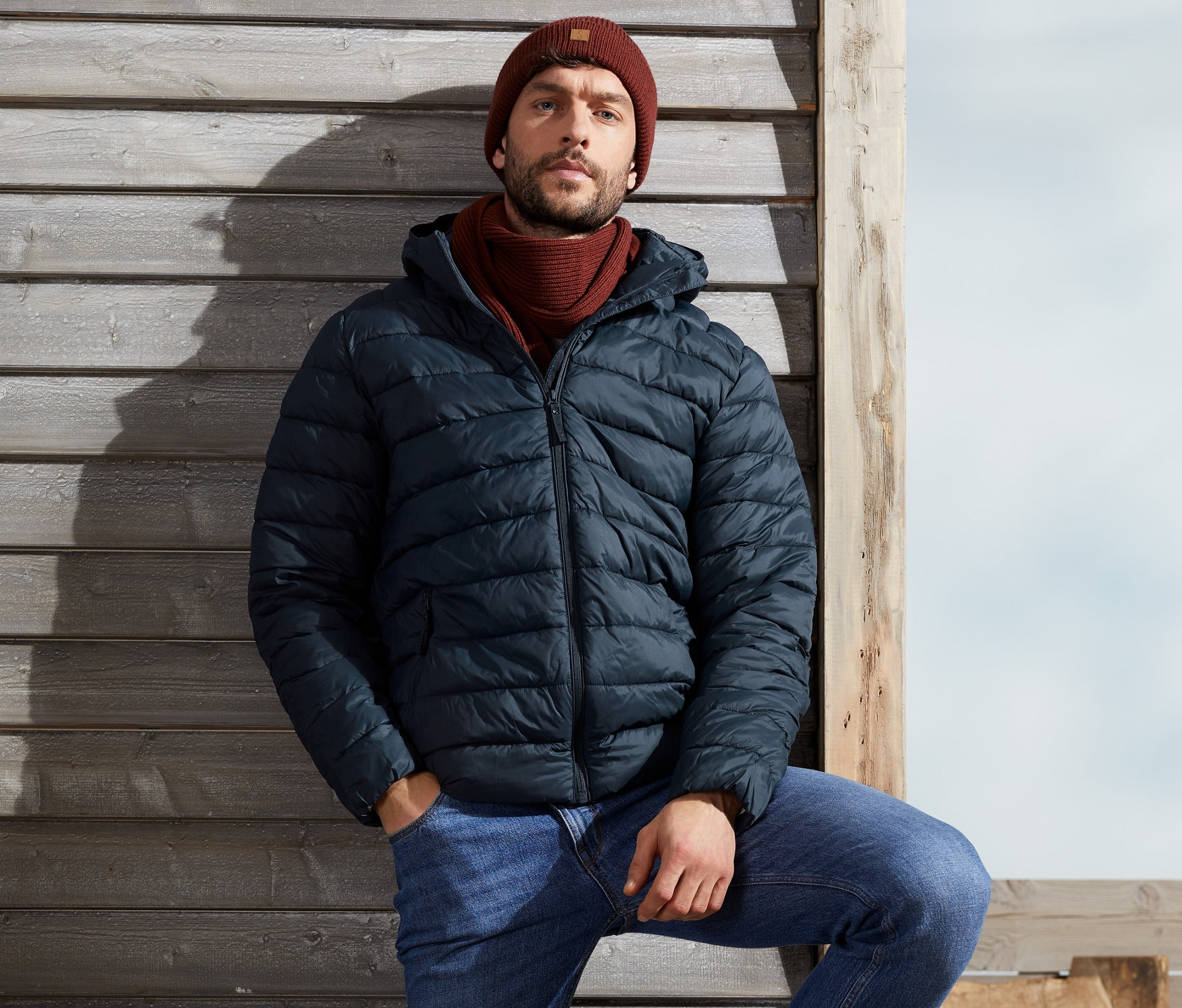 Mann lehnt an Holzwand und trägt eine blaue Steppjacke, Jeans und braune Stiefel.