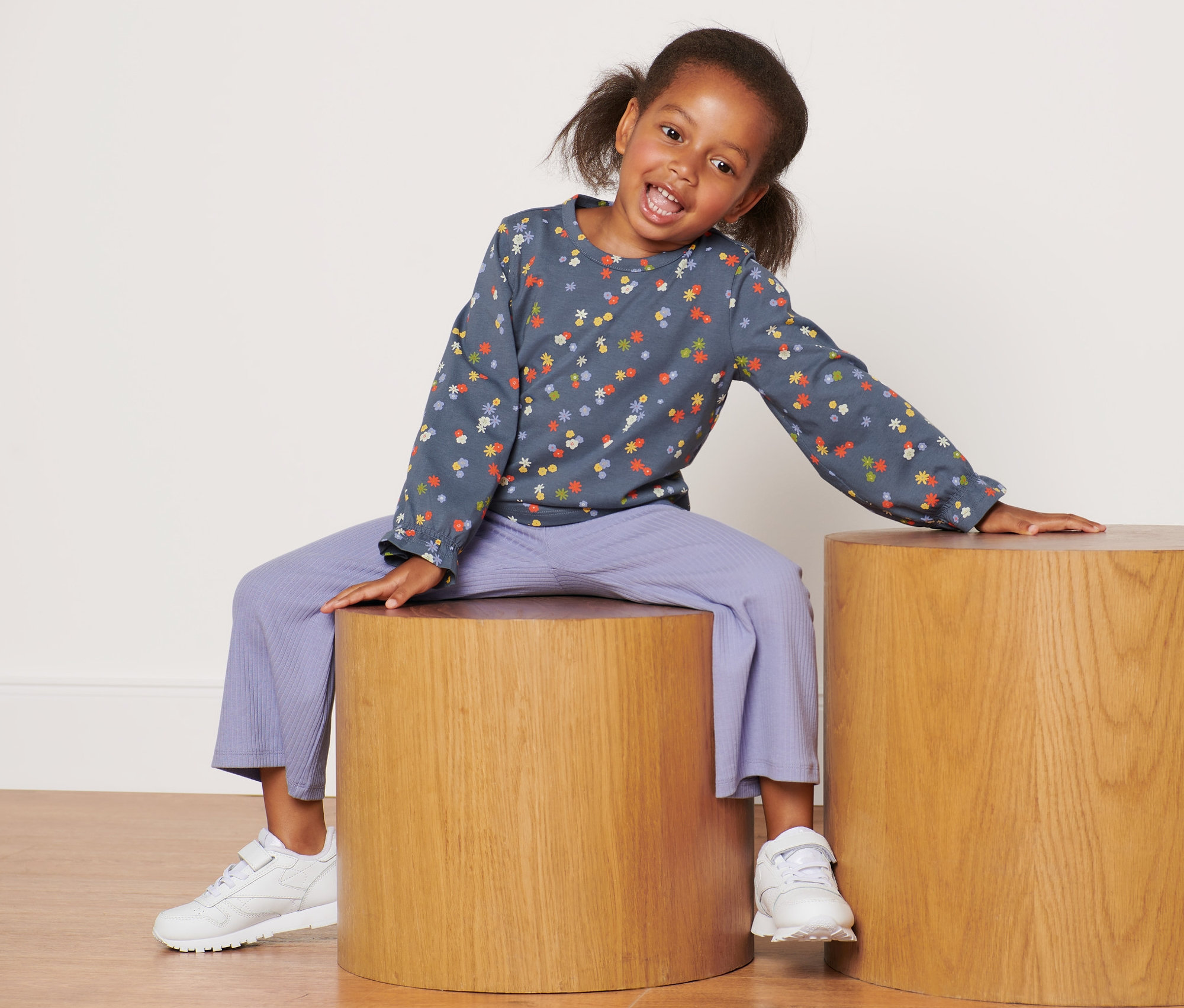 Ein lächelndes Mädchen sitzt auf einem Holzhocker und trägt ein Kleinkind-Langarmshirt.